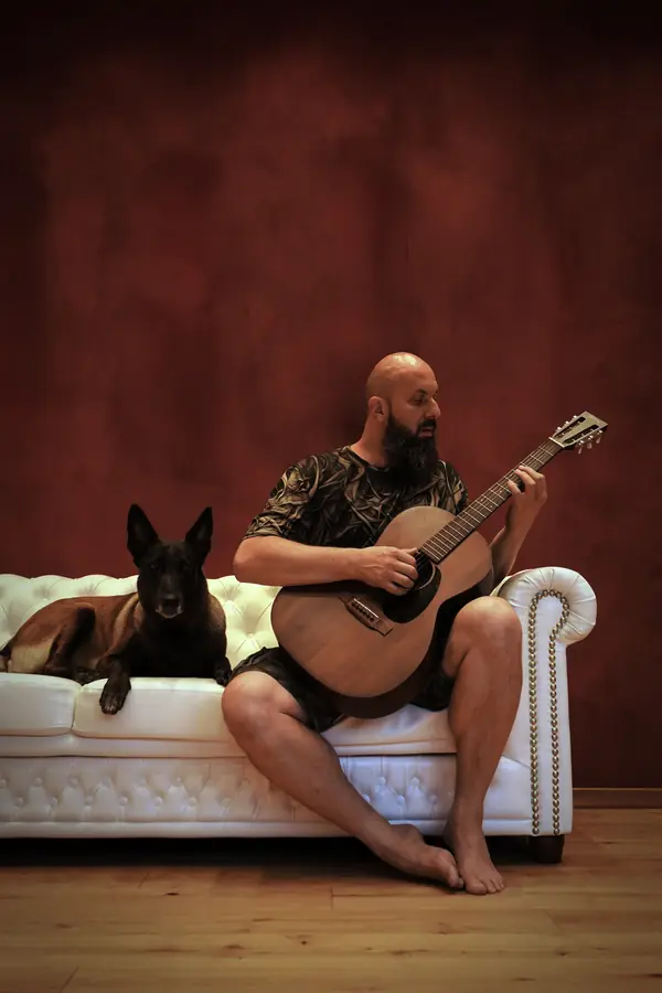 Rogue Bard on a white chesterfield, playing an acoustic guitar, his dog lying beside him